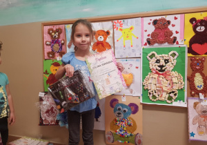 dziewczynka stoi przy wystawie z pracami plastycznymi z misiami trzyma w rękach nagrodę