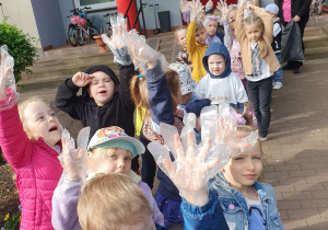 Grupa dzieci stoi przed przedszkolem parami podnoszą do góry rękę w plastikowej rękawiczce