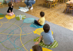 Dzieci siedząc na dywanie słuchają informacji przekazywanych o kundelku.