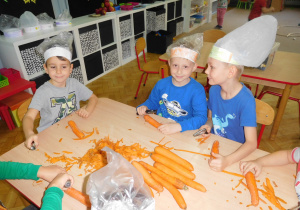 dzieci w czapkach kucharskich obierają marchew za pomocą skrobaków