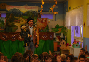 aktor z kukiełką na tle sceny teatralnej oraz widownia dzieci