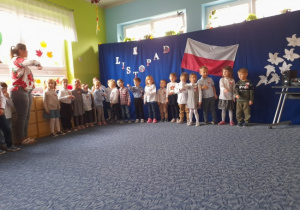 grupa dzieci ubrana w białe bluzki i ciemne spodnie lub spódniczki w sali przedszkolnej na tle flagi polski