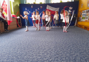 grupa dzieci ubrana w białe bluzki i ciemne spodnie lub spódniczki w sali przedszkolnej na tle flagi polski tańcząca ze wstążkami biało-czerwonymi