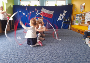 grupa dzieci ubrana w białe bluzki i ciemne spodnie lub spódniczki w sali przedszkolnej na tle flagi polski tańcząca ze wstążkami biało-czerwonymi
