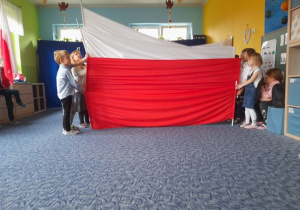 grupa dzieci ubrana w białe bluzki i ciemne spodnie lub spódniczki w sali przedszkolnej na tle flagi polski tańcząca ze suknem biało-czerwonymi