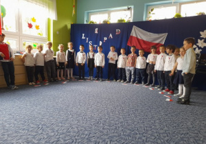 grupa dzieci ubrana w białe bluzki i ciemne spodnie lub spódniczki w sali przedszkolnej na tle flagi polski
