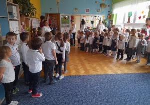 grupa dzieci ubrana w białe bluzki i ciemne spodnie lub spódniczki w sali przedszkolnej