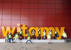 Grupa dzieci ubrana w kamizelki odblaskowe stojąca przy dużym napisie witamy.